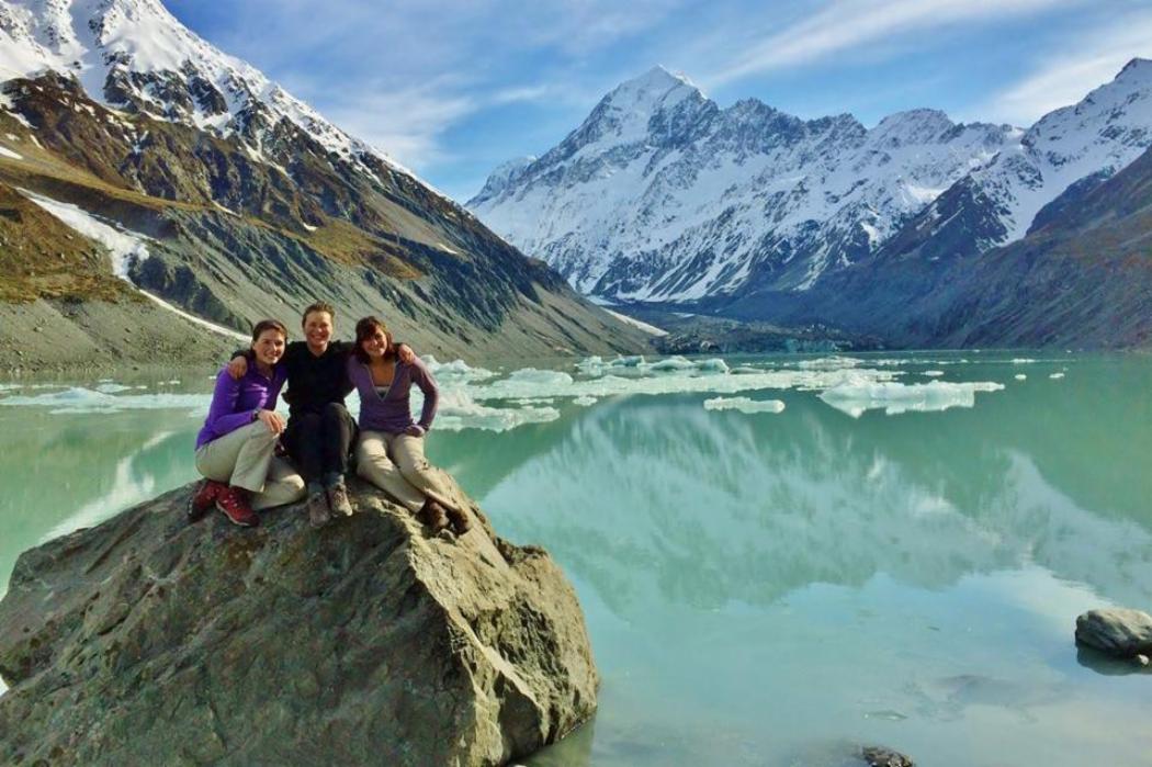 Study Abroad and Exchange Group of students sitting on rock together with a glacial lake and snowy mountain range behind them.